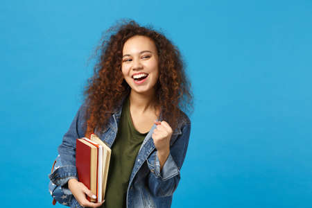 Young african american girl teen student in denim clothes, backpack hold books isolated on blue background studio portrait. Education in high school university college concept. Mock up copy spaceの写真素材