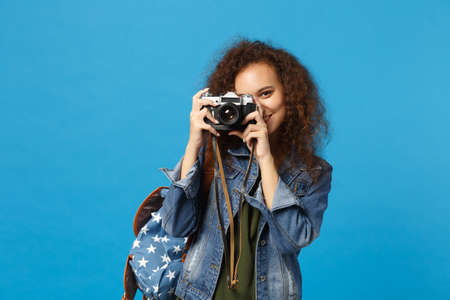 Young african american girl teen student in denim clothes backpack hold camera isolated on blue background studio portrait. Education in high school university college concept. Mock up copy spaceの写真素材