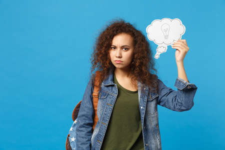 Young african american girl teen student in denim clothes, backpack hold clock isolated on blue...