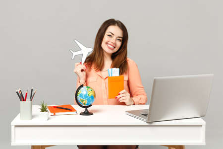 Young smiling woman holding paper air plane, Earth world globe, passport boarding pass ticket work at desk with pc laptop isolated on gray background. Achievement business career lifestyle conceptの写真素材