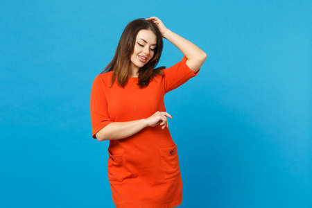 Beautiful brunette young woman wearing red orange dress looking aside standing posing isolated over trendy blue wall background, studio portrait. People lifestyle fashion concept. Mock up copy spaceの写真素材