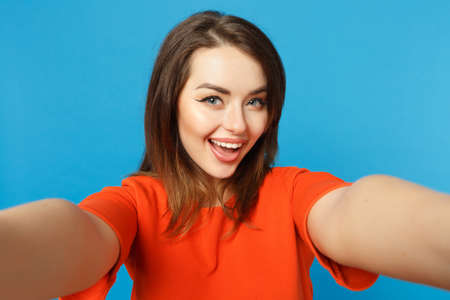 Close up photo of young woman in red orange clothes looking camera posing doing selfie shot isolated over trendy blue background, studio portrait. People lifestyle fashion concept. Mock up copy spaceの写真素材