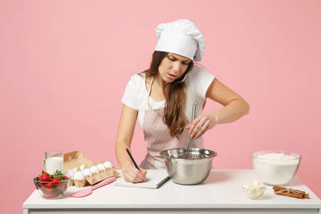 Chef cook or baker in white t-shirt, toque chefs hat cooking cake or cupcake at table, talking on cellphone, take an order whip dough in bowl with whisk isolated on pink pastel background in studioの写真素材