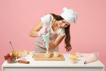 Cute housewife chef cook confectioner or baker in white t-shirt, toque chefs hat cooking at table isolated on pink pastel background in studio. Cupcake making process. Mock up copy space food conceptの写真素材
