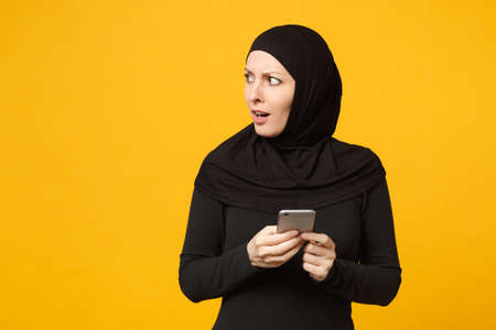 Smiling young arabian muslim woman in hijab black clothes hold in hands cellphone, chatting isolated on yellow wall background, studio portrait. People religious lifestyle concept. Mock up copy spaceの写真素材