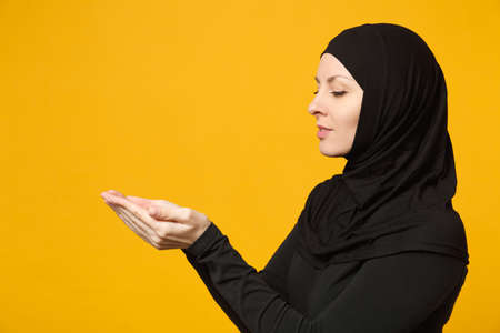 Side view young arabian muslim woman in hijab black clothes hold in hands empty workspace isolated on yellow wall background, studio portrait. People religious lifestyle concept. Mock up copy spaceの写真素材