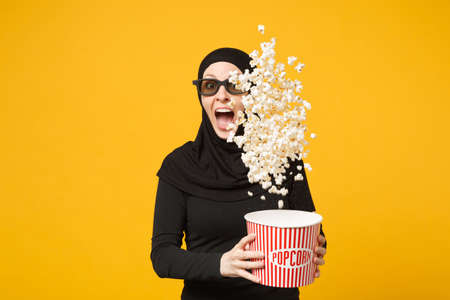 Muslim woman in hijab black clothes 3d glasses watch movie film hold popcorn, popping up pop corn isolated on yellow wall background studio portrait.の写真素材
