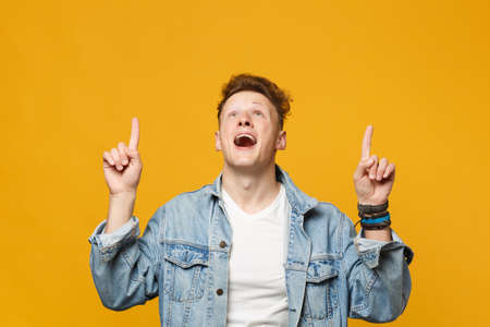 Amazed young man in denim casual clothes keeping mouth open pointing index finger up isolated on...