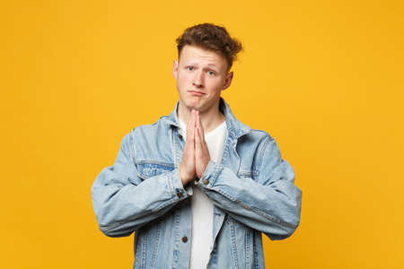 Portrait of pleading young man in denim casual clothes looking camera praying, holding hands folded...