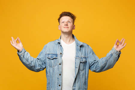 Handome young man in denim casual clothes looking up hold hands in yoga gesture relaxing meditating isolated on yellow orange background. People sincere emotions lifestyle concept. Mock up copy space