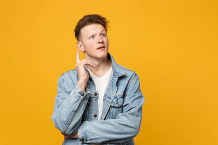 Pensive young man in denim casual clothes looking aside holding index finger up with great new idea isolated on yellow orange background. People sincere emotions lifestyle concept. Mock up copy spaceの写真素材