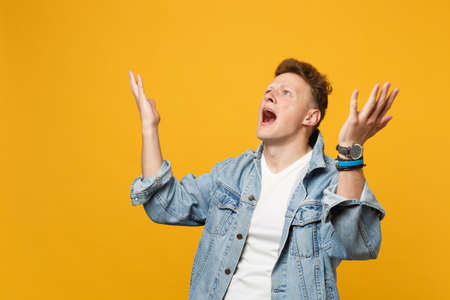 Irritated shocked young man in denim casual clothes looking up, spreading hands swearing isolated on yellow orange background in studio. People sincere emotions, lifestyle concept. Mock up copy spaceの写真素材
