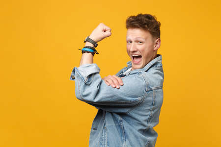 Side view of cheerful young man in denim casual clothes looking camera showing biceps, muscles...