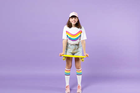 Portrait of joyful teen girl in vivid clothes standing, holding yellow skateboard isolated on violet pastel wall background in studio. People sincere emotions, lifestyle concept. Mock up copy spaceの写真素材