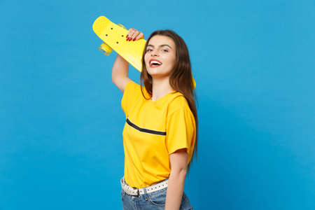 Portrait of laughing young woman in vivid casual clothes standing, looking camera, holding yellow skateboard isolated on blue wall background in studio. People lifestyle concept. Mock up copy spaceの写真素材