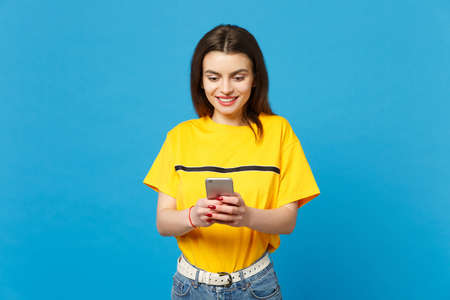 Portrait of smiling beautiful young woman in vivid casual clothes using mobile phone typing sms message isolated on bright blue wall background in studio. People lifestyle concept. Mock up copy spaceの写真素材