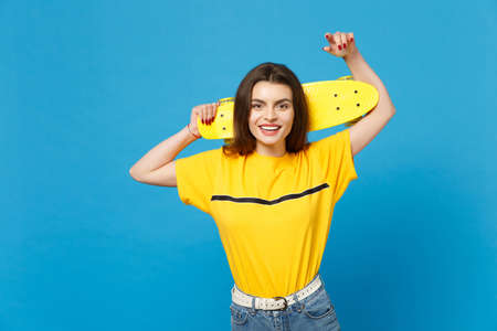 Portrait of smiling young woman in vivid casual clothes standing, looking camera, holding yellow skateboard isolated on blue wall background in studio. People lifestyle concept. Mock up copy spaceの写真素材