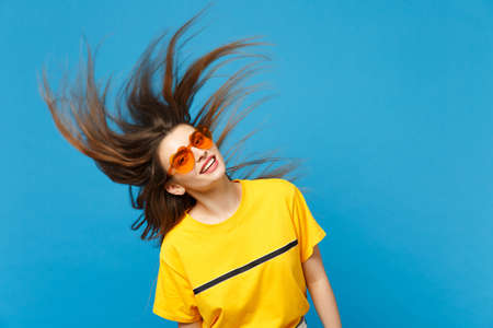 Portrait of pretty funny young woman in vivid casual clothes heart eyeglasses with flying hair, looking camera isolated on blue wall background in studio. People lifestyle concept. Mock up copy spaceの写真素材