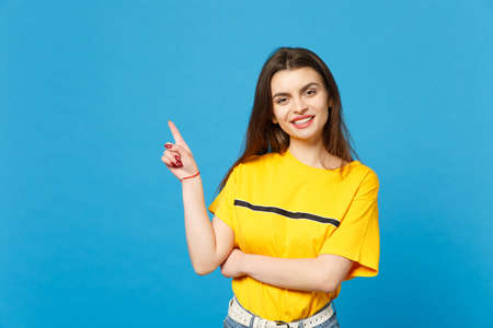Portrait of smiling beautiful young woman in vivid casual clothes looking camera, pointing index finger aside up isolated on bright blue wall background. People lifestyle concept. Mock up copy spaceの写真素材