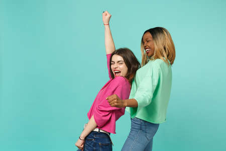 Two young women friends european and african american in pink green clothes dancing fooling isolated on blue turquoise wall background, studio portrait. People lifestyle concept. Mock up copy spaceの写真素材
