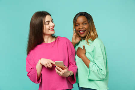 Two women friends european and african american in pink green clothes hold in hand cellphone...