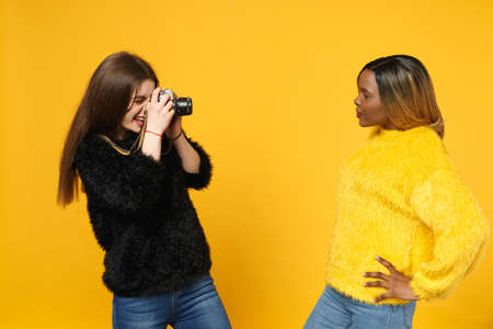 Two young women friends european and african american in black yellow clothes standing posing isolated on bright orange wall background, studio portrait. People lifestyle concept. Mock up copy spaceの写真素材