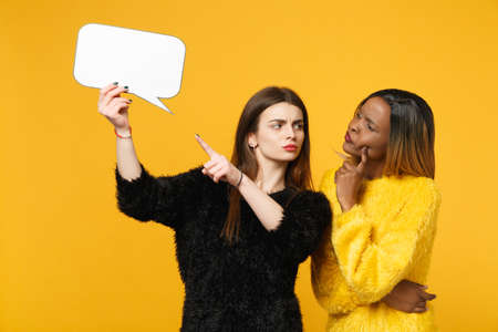 Two young women friends european and african american in black yellow clothes hold say clouds isolated on bright orange wall background, studio portrait. People lifestyle concept. Mock up copy spaceの写真素材