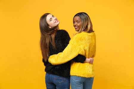Back view women friends european and african american in black yellow clothes standing posing isolated on bright orange wall background, studio portrait. People lifestyle concept. Mock up copy spaceの写真素材