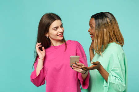 Two women friends european and african american in pink green clothes hold in hand cellphone isolated on blue turquoise wall background, studio portrait. People lifestyle concept. Mock up copy spaceの写真素材
