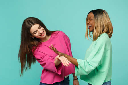 Two young women friends european and african american in pink green clothes standing posing isolated on blue turquoise wall background, studio portrait. People lifestyle concept. Mock up copy spaceの写真素材