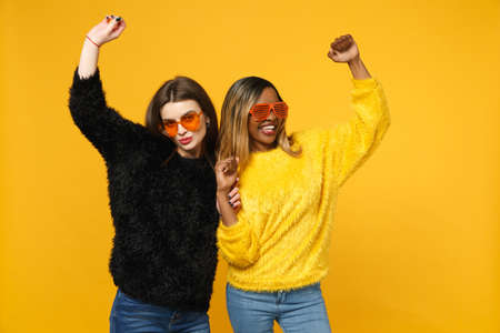 Two young women friends european and african american in black yellow clothes standing posing isolated on bright orange wall background, studio portrait. People lifestyle concept. Mock up copy spaceの写真素材