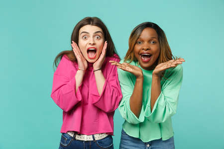 Two young shocked surprised women friends european and african american in pink green clothes isolated on blue turquoise wall background, studio portrait. People lifestyle concept. Mock up copy spaceの写真素材