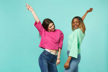 Two young women friends european and african american in pink green clothes dancing fooling isolated...