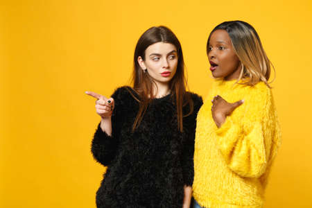 Two young women friends european and african american in black yellow clothes standing posing isolated on bright orange wall background, studio portrait. People lifestyle concept. Mock up copy spaceの写真素材