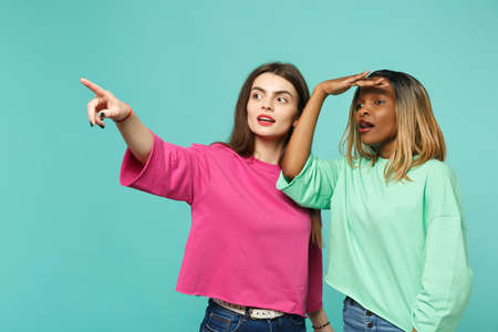 Two women friends european and african american in pink green clothes holding hand at forehead looking far away isolated on blue turquoise background. People lifestyle concept. Mock up copy space