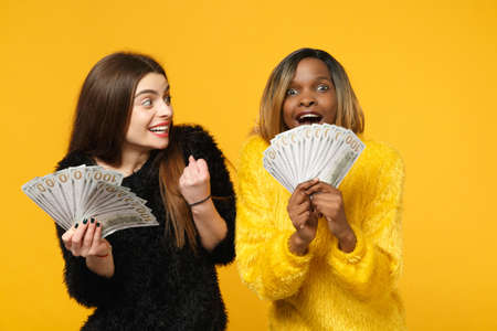 Two fun young women friends european and african in black yellow clothes holding fan of cash money in dollar banknotes isolated on orange wall background. People lifestyle concept. Mock up copy spaceの写真素材