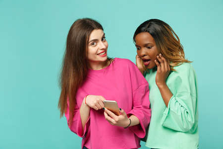 Two women friends european and african american in pink green clothes hold in hand cellphone isolated on blue turquoise wall background, studio portrait. People lifestyle concept. Mock up copy spaceの写真素材