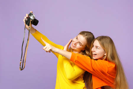 Two funny young blonde twins sisters girls in colorful clothes doing selfie shot on retro vintage photo camera isolated on violet blue background. People family lifestyle concept. Mock up copy spaceの写真素材