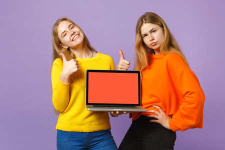 Two young blonde twins sisters girls showing thumb up holding laptop pc computer with blank empty screen isolated on pastel violet blue background. People family lifestyle concept. Mock up copy spaceの写真素材