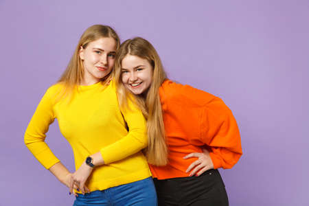 Two pretty smiling young blonde twins sisters girls in vivid colorful clothes standing, looking camera isolated on pastel violet blue background. People family lifestyle concept. Mock up copy spaceの写真素材