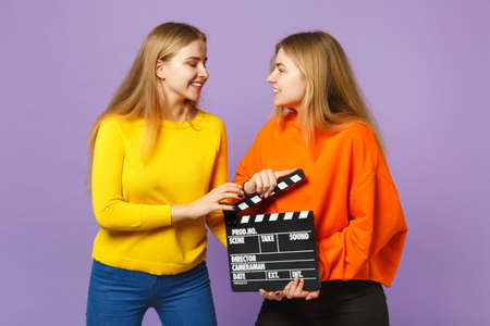 Two smiling young blonde twins sisters girls in colorful clothes holding classic black film making clapperboard isolated on violet blue background. People family lifestyle concept. Mock up copy spaceの写真素材