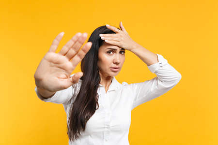 Portrait of concerned young woman in white shirt putting hand on head showing stop gesture with palm isolated on yellow orange wall background in studio. People lifestyle concept. Mock up copy spaceの写真素材