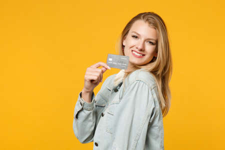 Portrait of smiling beautiful young woman in denim casual clothes looking camera holding credit bank card isolated on yellow orange background in studio. People lifestyle concept. Mock up copy spaceの写真素材