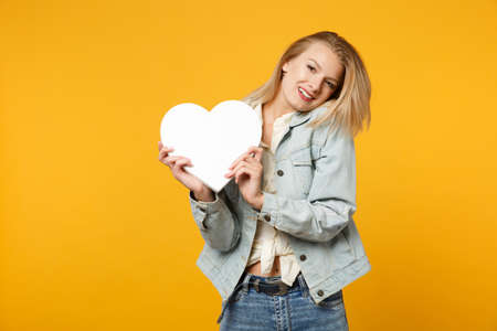 Portrait of pretty young woman in denim casual clothes looking camera holding white heart with copy space isolated on yellow orange background in studio. People lifestyle concept. Mock up copy spaceの写真素材