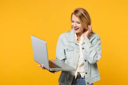 Portrait of laughing joyful young woman in denim casual clothes holding, using laptop pc computer isolated on yellow orange wall background in studio. People lifestyle concept. Mock up copy spaceの写真素材
