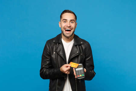 Young unshaven man black jacket white t-shirt hold in hand wireless modern bank payment terminal to process and acquire credit card payments isolated on blue wall background. People lifestyle conceptの写真素材