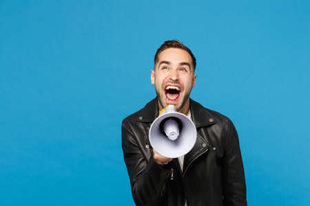 Handsome young unshaven man in black jacket white t-shirt scream in megaphone, announces discounts sale isolated on blue wall background studio portrait. People lifestyle concept. Mock up copy spaceの写真素材