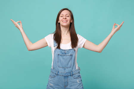 Smiling young woman girl in casual denim clothes isolated on blue turquoise background. People lifestyle concept. Mock up copy space. Hold hands in yoga gesture, relax meditating, keeping eyes closed.の写真素材