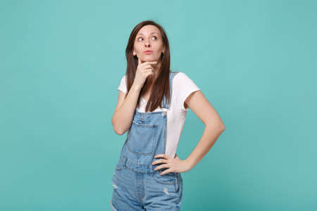 Pensive young brunette woman girl in casual denim clothes posing isolated on blue turquoise background in studio. People lifestyle concept. Mock up copy space. Put hand prop up on chin, looking up.の写真素材