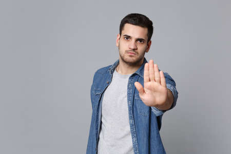 Angry mad sad upset unshaven young man in denim jeans shirt posing isolated on grey wall background studio portrait. People sincere emotions lifestyle concept. Mock up copy space. Stop palm gesturing.の写真素材